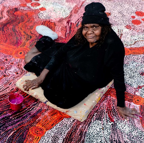 Aboriginal woman dressed in black sitting on painted artwork of red, purple and white