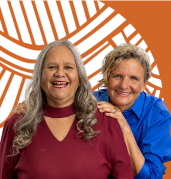 Aboriginal Elder Aunty Munya and ally Carol rogers with a orange and white design in the background. Aunty Munya is wearing a red top and Carla standing behind in a blue top. Both are smiling.