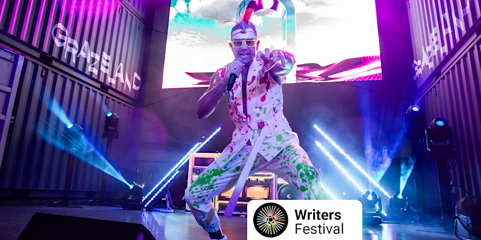 Image of performer on a stage with bright lights, wearing a paint splattered white outfit, feather headband and yellow glasses