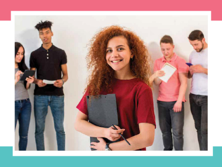 Image of 5 young people, 4 in the background and 1 standing at the front with red hair, a red t-shirt holding a document book.